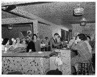Vintage press photo: MIAMI, FLA: DEC-00-1940: The patience of Job, the friendliness of hundreds of delivery truck drivers as collectors and contributors has enabled Joe Wiser and his wife to build “Bottle Cap Inn” a bar in Miami at 1290 N. 119th St., whose walls and ceilings are decorated with over a million bottle caps. It took three years, a ton of nails and a ton of cement to do the job. Bottle caps are everywhere, on the floor, ceiling, bar, clock, lights, chairs, cash register, curtains, and even on the driveway. It’s the most unique rendezvous in Florida. Caps have been sent by admirers from the four corners of the world. Bottle tops, over the bar, and under it too, in the table tops, the ceiling and everywhere. Here’s the proud proprietor serving his guest. This photo gratis for the City of Miami. Miami New Service, Hamilton Wright Jr., Executive Editor.